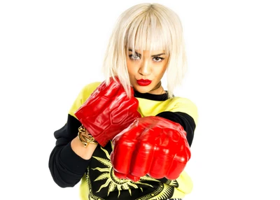 Rita Ora posa com confiança, exibindo luvas de boxe vermelhas oversized, estilizada em um traje vibrante com um lábio ousado, incorporando uma fusão de moda e atletismo.