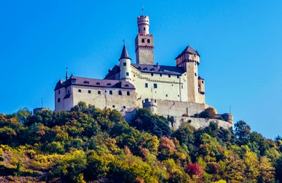 Un majestueux château médiéval perché au sommet d'une colline, entouré d'un feuillage automnal vibrant et d'un ciel bleu clair, mettant en valeur ses impressionnantes tourelles et fortifications comme un monument historique emblématique.