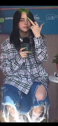 Uma pessoa com cabelo tingido de verde, vestindo uma camisa oversized xadrez e shorts de denim desgastados, posa com confiança enquanto faz um gesto brincalhão.