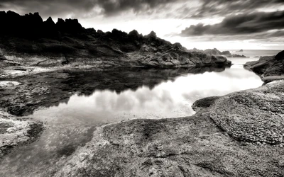 Costa monocromática con formaciones rocosas y agua reflectante bajo nubes dramáticas.