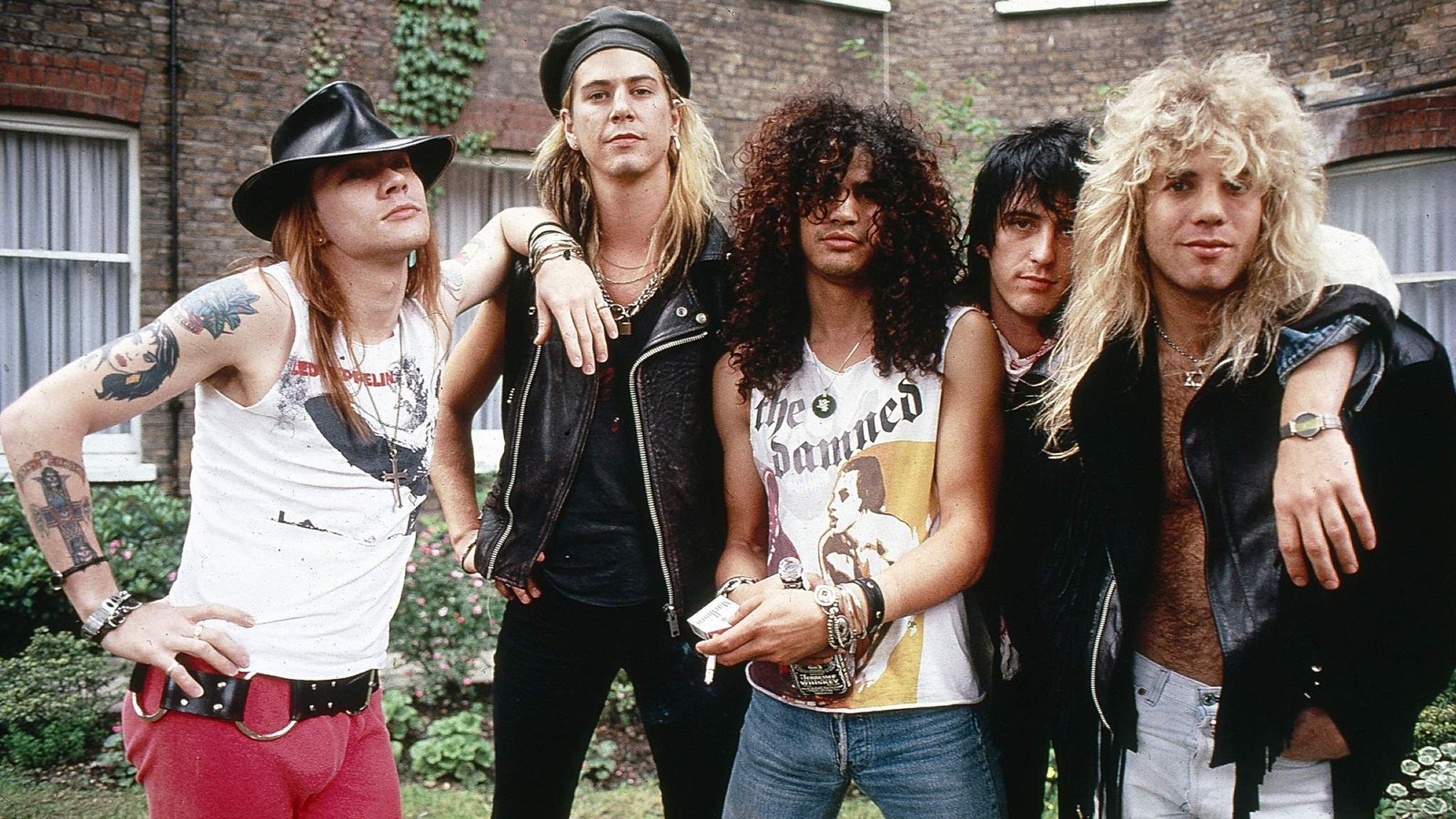 Un grupo arqueado de jóvenes de pie frente a un edificio de ladrillo (axl rose, slash, duff mckagan, izzy stradlin, guns n roses)