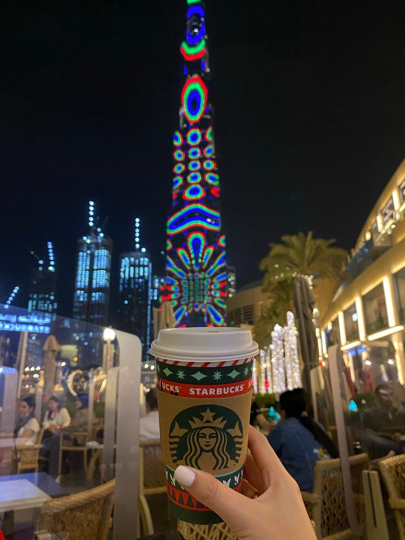 Чашка starbucks в руке на фоне городского горизонта (дубай, dubai, burj khalifa, освещение, рождественские огни)