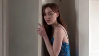 Phoebe Dynevor em um vestido de renda azul, capturada em um momento espontâneo perto de uma porta.