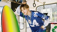 Jeno do NCT Dream posando de forma brincalhona com uma prancha de surf em um cenário de ônibus vintage