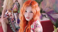 Minatozaki Sana from TWICE with vibrant orange hair, exuding a playful and captivating charm against a colorful backdrop.