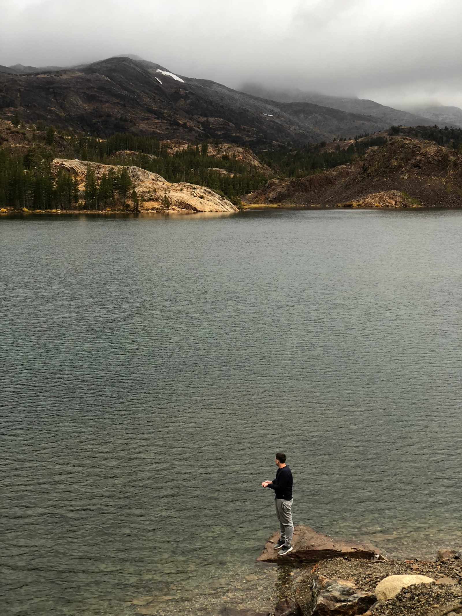 There is a man standing on a rock by the water (water, lake district, mountainous landforms, water resources, highland)