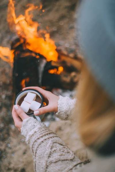 Gemütliche Hände halten heiße Schokolade mit Marshmallows vor einem warmen Feuer.