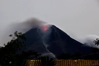 Monte Merapi em erupção: Um vulcão escudo emitindo lava e cinzas contra um céu nublado.