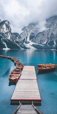 Serene lake surrounded by majestic mountains, dotted with wooden boats along a scenic dock under a cloudy sky.