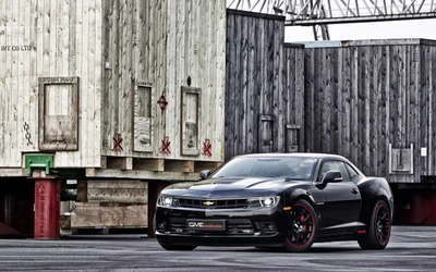 Chevrolet Camaro SS preto estacionado contra um fundo de caixas industriais, exibindo seu design elegante e características focadas em desempenho.