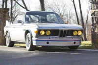 Classique BMW E9 Coupé avec des phares jaunes distinctifs et des bandes de course, sur fond de paysage pittoresque.