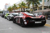 Uma fila de carros esportivos exóticos, incluindo um Mazda RX-7 personalizado em destaque e um impressionante Lamborghini Aventador, estacionados ao longo de uma rua ladeada de palmeiras, exibindo designs vibrantes e tuning automotivo.