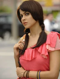 A young woman with long, dark hair styled in pigtails, wearing a pink ruffled top and colorful bangles, gazes thoughtfully into the distance.