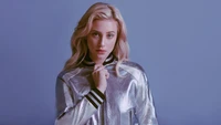 Lili Reinhart dans une veste bomber argentée élégante sur un fond bleu doux.