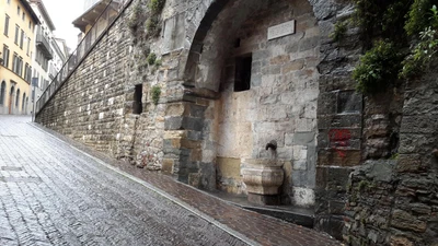 Mittelalterliche Steinmauer mit einem Bogen und einem Wasserbrunnen, gelegen in einer Kopfsteinpflastergasse in Mailand, die den historischen Charme der Stadt widerspiegelt.