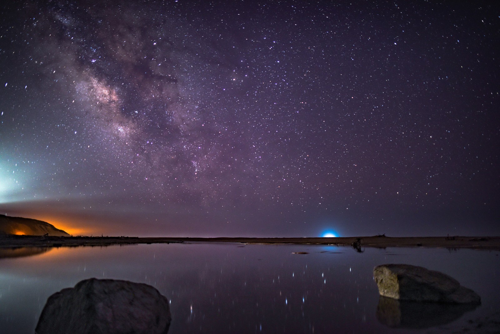 Uma vista de um lago com pedras e uma luz azul brilhante à distância (nuvem, reflexo, estrelas, céu noturno, natureza)