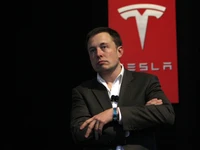 Elon Musk delivering a speech on Tesla's innovations, with a prominent Tesla logo in the background.