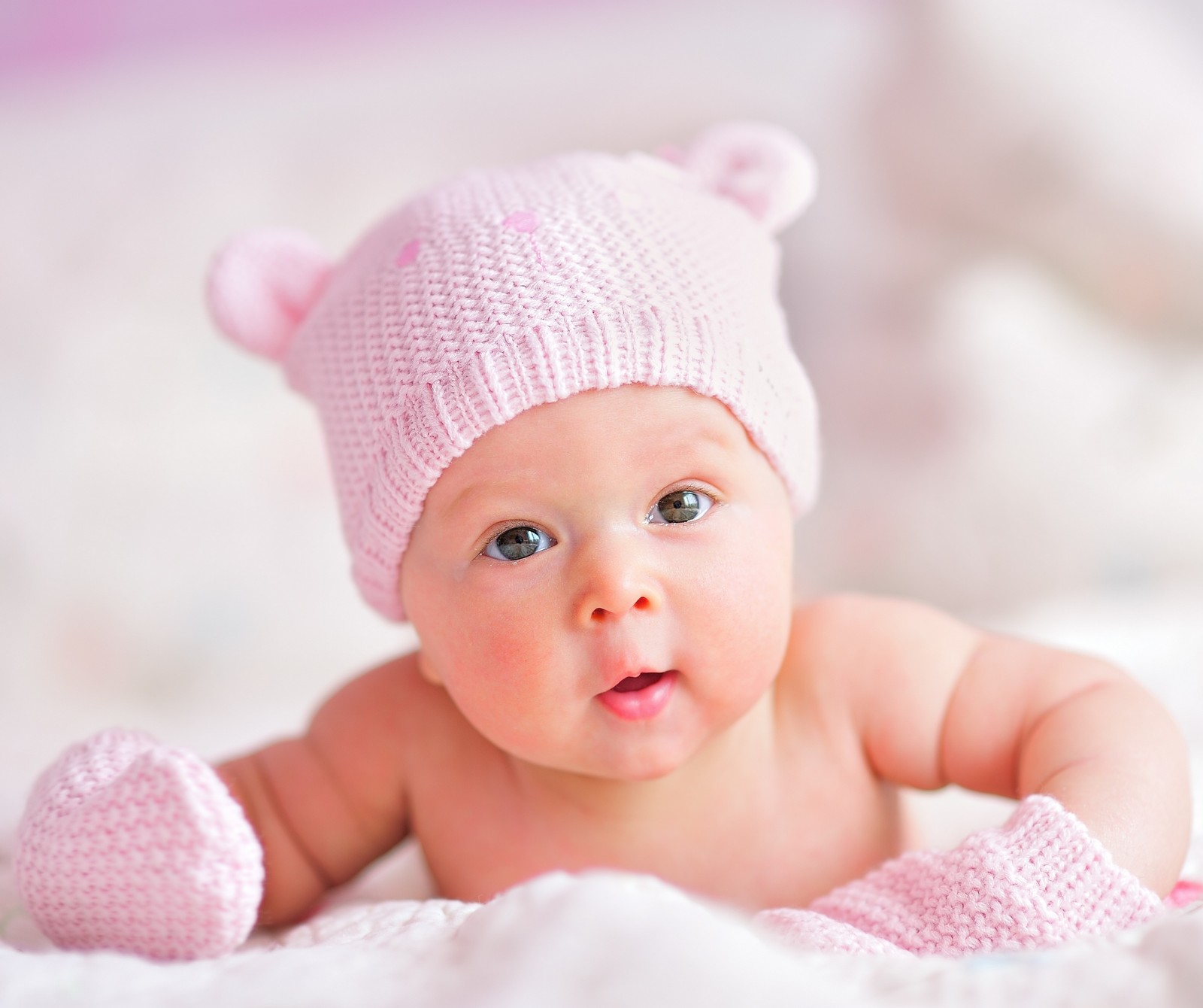 There is a baby wearing a pink hat and mittens on a bed (meem, rubab)