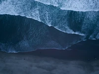 Vista aérea de las olas rompiendo en una playa de arena oscura, mostrando el contraste entre las aguas azules del océano y la costa.