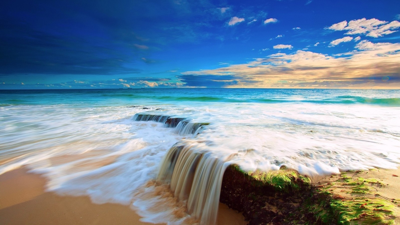 A view of a beach with a small waterfall coming out of the water (ocean, wave, sea, shore, wind wave)