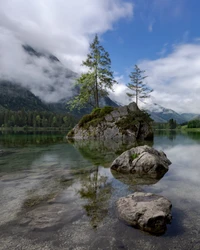 Cena de lago tranquilo com afloramentos rochosos e árvores refletindo em águas calmas, emoldurada por montanhas e nuvens.