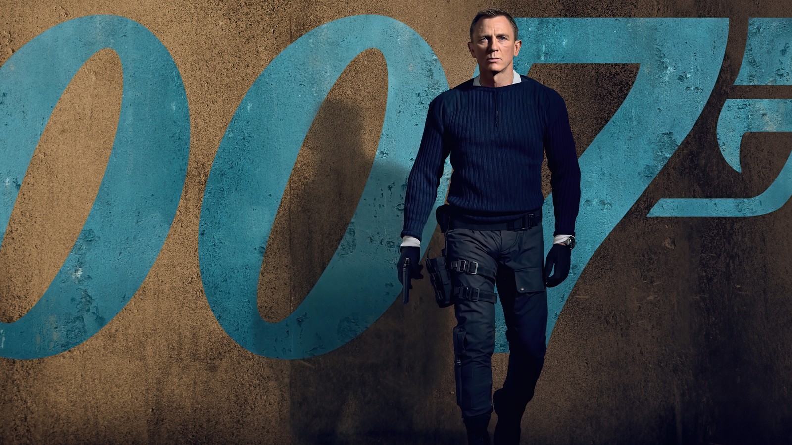 Un hombre en un suéter azul y pantalones negros de pie frente a una pared con un logotipo azul (no time to die, película, 007, james bond, daniel craig)