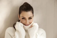 Kim Kardashian in einem gemütlichen Pullover, lächelt warm vor einem strukturierten Hintergrund.