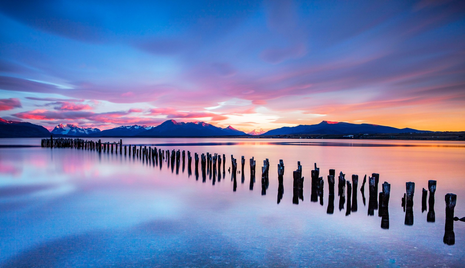 Uma foto de longa exposição de um pôr do sol sobre um lago com postes de madeira (parque nacional torres del paine, torres del paine national park, chile, por do sol, cenário)