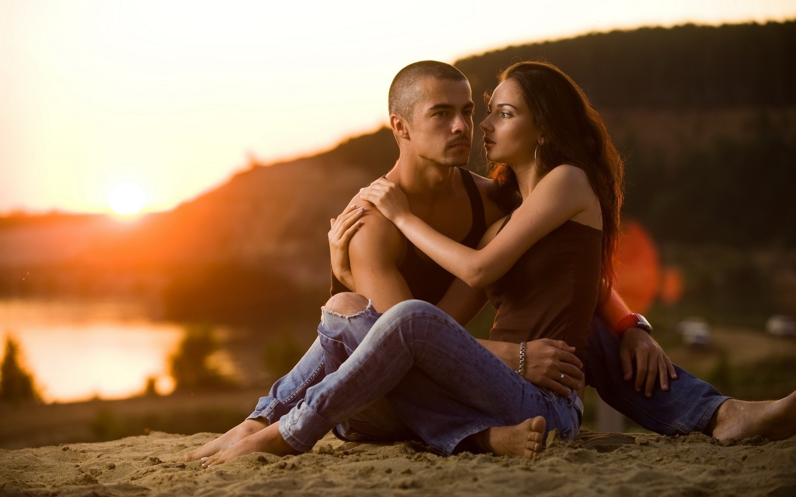 Um casal sentado na praia ao pôr do sol com um lago ao fundo (abraço, romance, beijo, casal, amor)