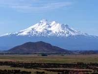 Majestic snow-capped stratovolcano rising above a lush, green landscape under a clear blue sky.