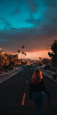 Atardecer sereno en Los Ángeles: una mujer pasea por una carretera flanqueada de palmeras bajo un cielo que brilla en naranja