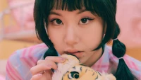 Chaeyoung de TWICE sostiene un tigre de peluche cerca, exudando un encanto juguetón pero cautivador contra un vibrante fondo rosa.