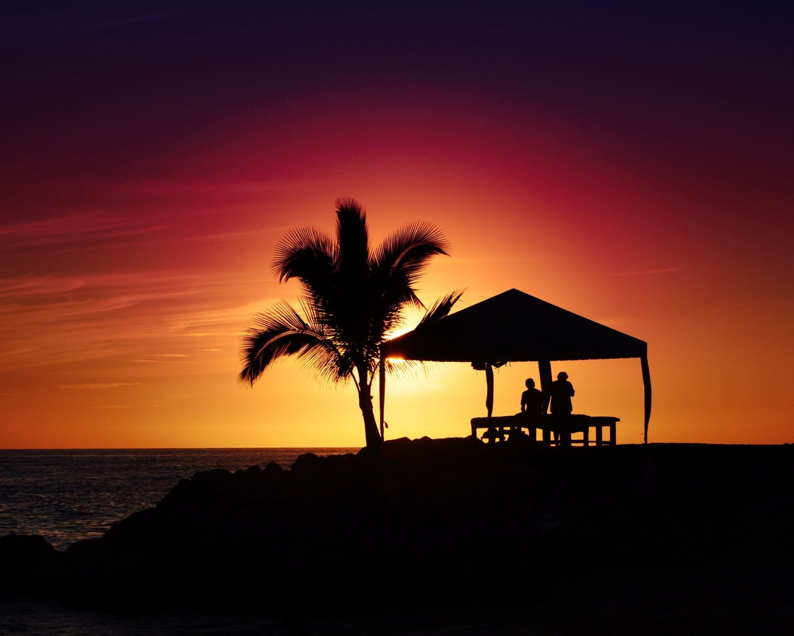 Pôr do sol na praia com um casal de pessoas sentadas sob um gazebo (praia, feriado, amor, por do sol)