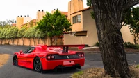 Ferrari F50 parked on a scenic street, showcasing its sleek design and vibrant red color against a backdrop of trees and adobe-style architecture.