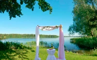 Un entorno sereno junto al lago con un arco decorativo cubierto de tela, rodeado de exuberante vegetación y un cuerpo de agua tranquilo que refleja el claro cielo azul. Ideal para una celebración de verano o recreación en la naturaleza.