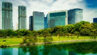 Eine ruhige Stadtlandschaft mit Wolkenkratzern und modernen Eigentumswohnungen vor einer üppig grünen Landschaft, die sich in ruhigem Wasser unter einem Tageshimmel in Tokio spiegelt.