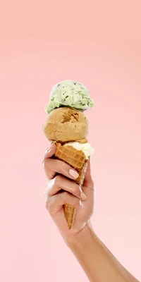 Cornet de glace à trois boules tenu dans une main, présentant des saveurs de vanille, caramel et menthe chocolatée sur un fond rose doux.