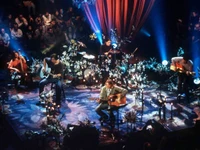 Nirvana's intimate acoustic performance surrounded by a floral stage setup, featuring musicians playing guitar and drums amidst an engaged audience.