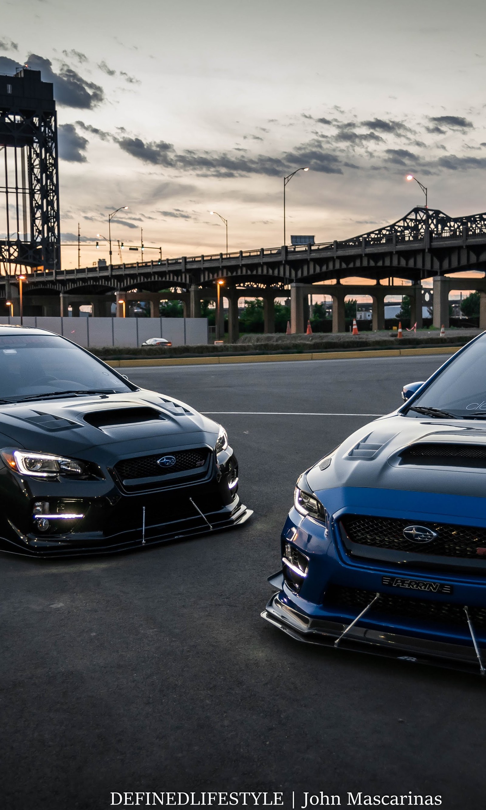 Dois carros subaru estacionados em um estacionamento com uma ponte ao fundo (carro, carros)