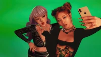 Ningning d'Aespa et un personnage virtuel posent pour un selfie sur un fond vert vif, montrant un mélange de mode moderne et d'esthétique K-pop.