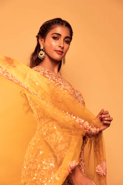 Pooja Hegde dans une magnifique tenue jaune ornée avec des boucles d'oreilles élégantes sur un fond chaud.