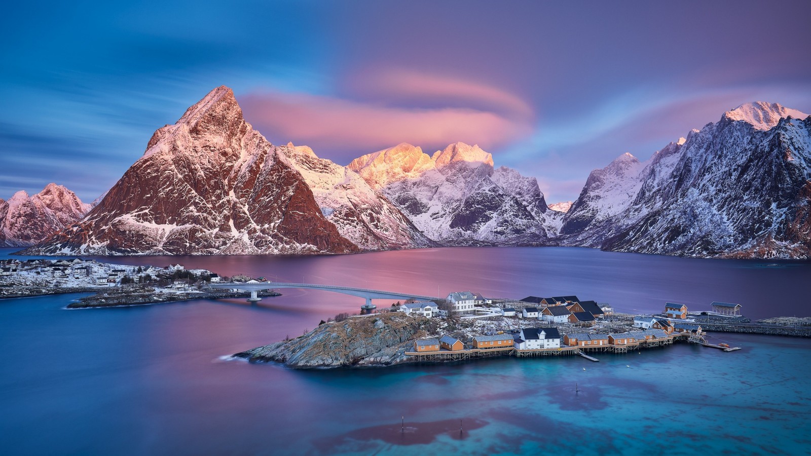 Una pequeña aldea en medio de un cuerpo de agua con montañas al fondo (lofoten, montaña, naturaleza, agua, nube)