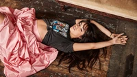 Camila Cabello in einer stilvollen Pose, trägt ein grafisches T-Shirt und einen fließenden rosa Rock, auf einem rustikalen Holzboden.