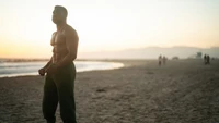 Jonathan Majors em um momento contemplativo na praia ao pôr do sol, incorporando força e introspecção.