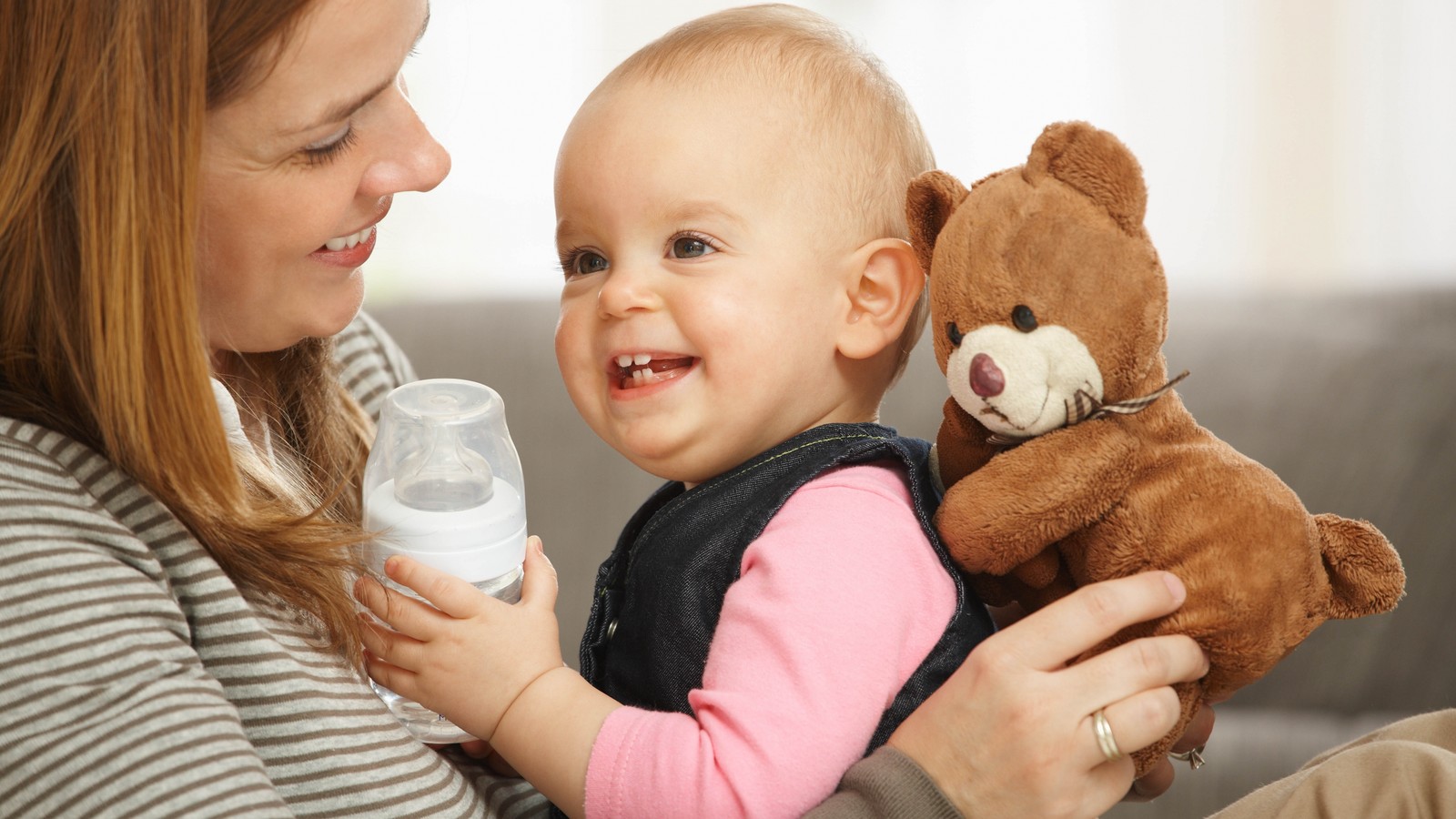 Arafed baby holding a bottle and a teddy bear while sitting on a couch (child, stuffed toy, girl, ear, child care)