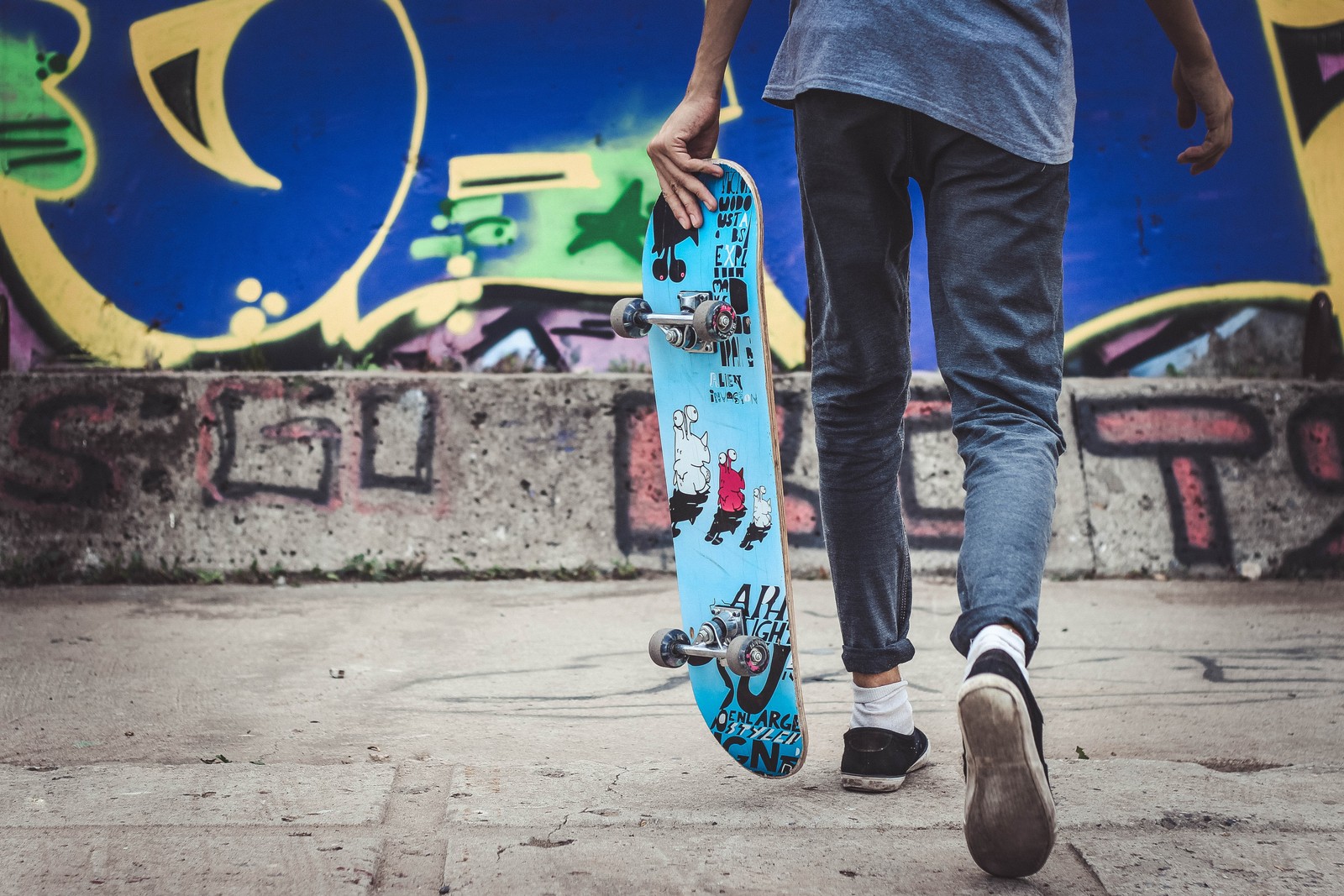 Arafed man walking with a skateboard in front of a graffiti covered wall (street fashion, skatepark, sports equipment, human leg, shoe)