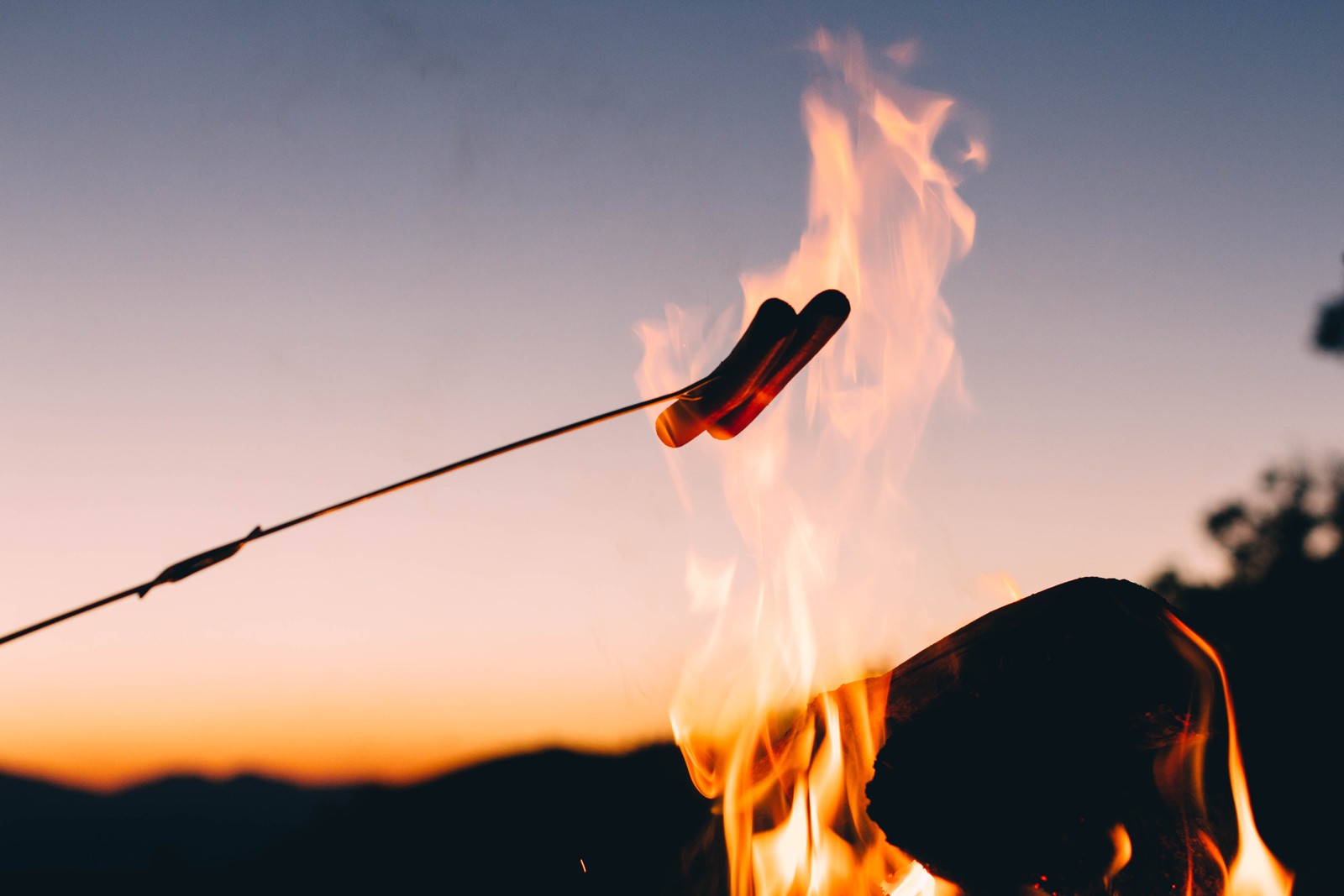 There is a hot dog on a stick being cooked over a campfire (campfire, heat, flame, fire, cloud)