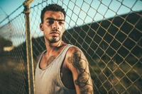 A muscular man with tattoos leans against a chain-link fence, exuding a relaxed yet confident demeanor in a sleeveless shirt.