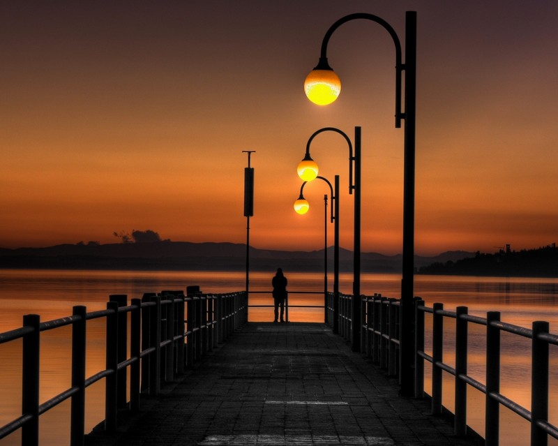 Une vue d'une jetée avec des lampadaires et une personne marchant dessus (soir, coucher de soleil)