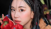 Chaeyoung do TWICE, com cabelo azul vibrante e uma expressão suave, segurando morangos frescos contra um fundo escuro.